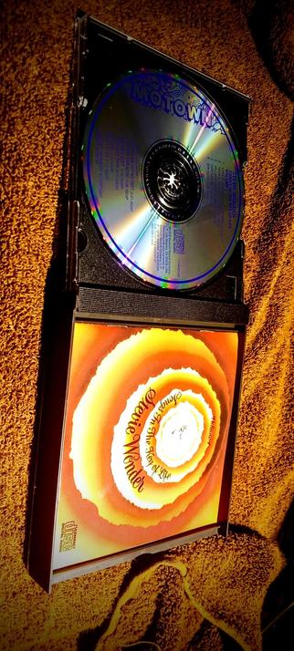 The composition shows a CD case with a CD inside, placed on a textured, orange-brown carpet. The CD case is open, revealing a CD with a reflective surface and text. The CD is labeled "MOTOWN" at the top and "STEVE WONDER VOLUME 1 SONGS IN THE KEY OF LIFE" in the center. The text on the CD includes track listings and other information. The case cover features a circular design with concentric rings in shades of orange, yellow, and white, with the text "STEVE WONDER SONGS IN THE KEY OF LIFE" in a stylized font. The bottom left corner of the case cover has the text "COMPACT DISC DIGITAL AUDIO." The overall lighting casts a warm glow on the scene, highlighting the CD and case against the carpet background.