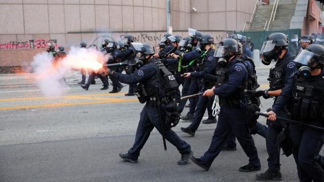 A line of black-uniformed Imperial Stormtroopers advance on the rebels in a scene from Andor, a sci-fi show that is part of the Star Wars franchise.

Wait, no. It's a line of heavily-armed riot police advancing on protesters in Los Angeles, June 2025. All the stormtroopers carry long arms (rifles, etc). One is in the act of firing a shot, creating a large flash from the muzzle of his weapon.

In the background are two pieces of graffiti on a concrete wall. One reads "Leviticus 19.33-34". The other, "Pew pew".