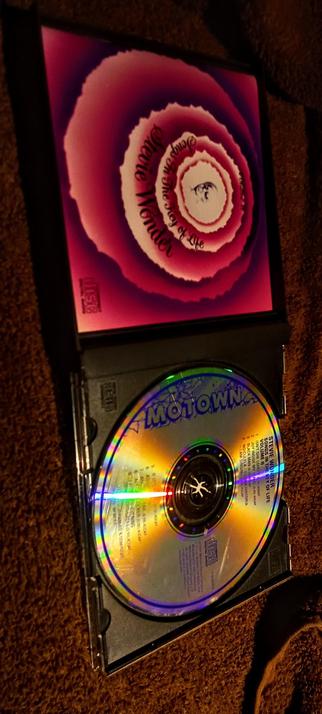 The photograph shows a CD case with a CD inside. The CD case is open, revealing a CD with a reflective surface that displays a rainbow-like pattern. The CD is labeled "Motown" at the top, with the text "STEVE WONDER SONGS IN THE KEY OF LIFE VOLUME II" on the right side. The case cover features a colorful, concentric circle design with a gradient of pink and purple hues, and the text "The Joy of Life" is visible in the center. The background of the image is a textured brown surface, likely a carpet. The CD case is positioned on the carpet, with the CD partially inserted into the case.

 Ovis2-8B

🌱 Energy used: 0.175 Wh