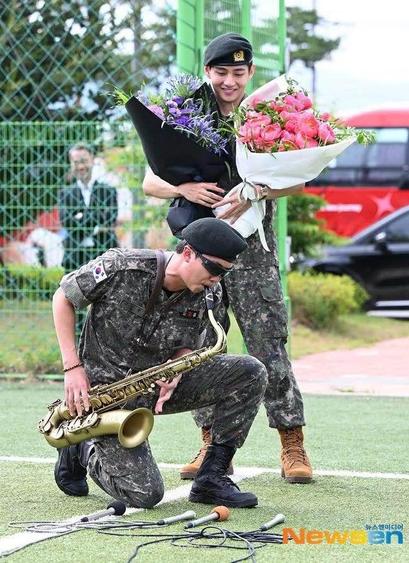 V of BTS carries two bouquets of flowers standing next to RM who is playing saxophone and kneeling on the ground.