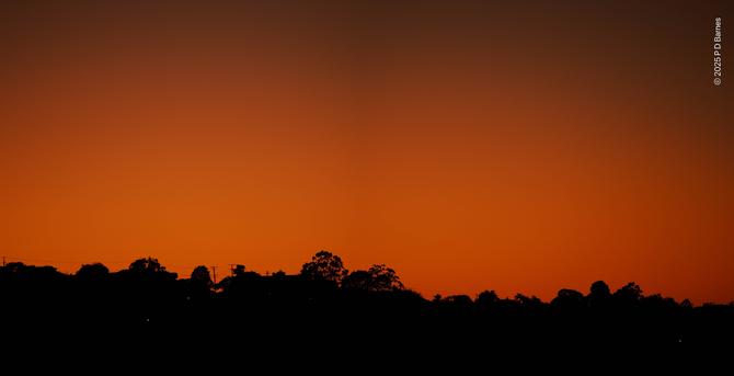 A clear, cloudless sky has begun to brighten into day, glowing a deep, burnt orange. It silhouettes a section of suburban ridgeline, still dark, showing mostly trees with an occasional roof and utility pole.