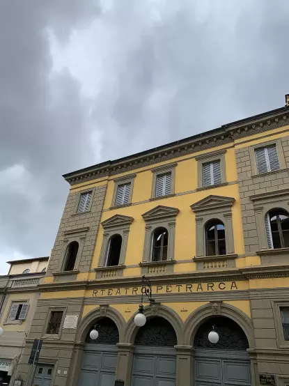 The image shows a building with a classical architectural style, featuring a yellow facade and large windows. The structure appears to be a theater, as indicated by the name "Teatro Petrarca" displayed above the entrance. The sky above is cloudy, suggesting overcast weather.

Image Credits: Wikimedia / Photo2023 / CC BY 4.0