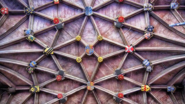 An intricately carved wooden vaulted ceiling features a repeating star-like pattern of crisscrossing beams. At the intersection points and within the geometric shapes, numerous colourful heraldic shields are embedded. The central shield is larger and more ornate, displaying a royal coat of arms. The wood has a rich, aged patina, with visible grain and variations in tone.