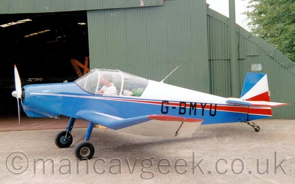 Side view of a single-engined light aircraft parked facing to the left on a black tarmac apron.
The plane has a white upper rear fuselage, with most of the rest being blue, with an additional red stripe running along the body and getting wider as it moves rearwards.
The black registration "G-BMYU" is overlaid on the blue and red sections on the rear fuselage.
2 people can be seen in the cockpit, with a load of bags visible in the rear of the cabin.
Grey concrete apron fills the foreground, with a green arched hangar with its doors slid wide open in the background, under bright but hazy skies.