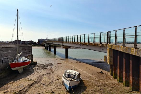 Photo of a 214 metre long footbridge with a pivoting 50 metre central span. Steel on concrete piles with laminated-glass parapets. For pedestrians and cyclists crossing the river Adur.  Low tide and there are a couple of boats in the foreground on the sand. The bridge is formally known as the Adur Ferry Bridge. Shoreham by Sea, West Sussex, UK.