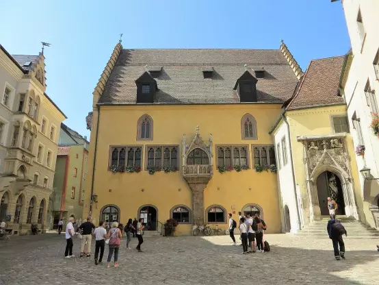 The image shows a building with a distinctive architectural style, featuring a mixture of medieval and Renaissance elements. It has a yellow facade, large windows, and decorative detailing. In front of the building, there are several people gathered, some taking photos and others walking. The setting appears to be a square or courtyard, surrounded by other structures, and the sky is clear and blue.

Image Credits: Wikimedia / Bärwinkel,Klaus / CC BY-SA 4.0