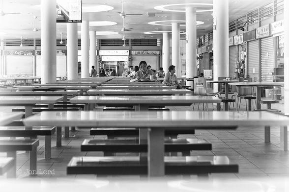 This Is A Black And White Indoor Street Photo In Landscape Format. In The Immediate Foreground Is A Rectangular Table With A Connected Bench Seat On Either Long Side. Moving Away From The Camera And Also In Parallel On The Left Is A Line Of Tables Continuing Into The Distance. The Tables Closest And Furthest From The Camera Are Blurred Out Of Focus. Halfway Along (Six Tables Away) Is A Young Chinese Woman Facing The Camera, Sitting And Eating From A Bowl With Chopsticks. She Is The Focal Point. Either Side Of The Tables Are Two Parallel Rows Of Round Columns Supporting The Roof. Between The Columns Is A Line Of Four Ceiling Fans With Two Further Lines Of Fans To The Left. Stretching Along The Right Margin And In The Background Are The Fronts Of Numerous Food Vendors, Most Are Shuttered Closed. 
The Location Is The Food Court (Hawker Centre) At The Tiong Bahru Market In Singapore (2017).