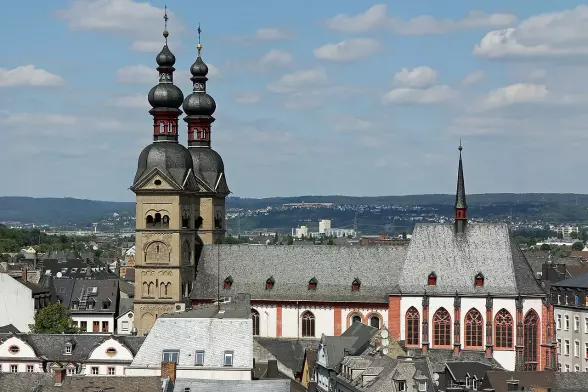 The image depicts a cityscape featuring a large building with distinctive architectural elements, including two prominent towers adorned with onion domes and a smaller spire. This building appears to be a church or cathedral, typical of European architecture, with detailed windows and a slate roof. Surrounding it are more buildings, set against a backdrop of hills and a cloudy sky. The overall scene suggests a historic urban area.

Image Credits: Wikimedia / Holger Weinandt / CC BY-SA 3.0 de