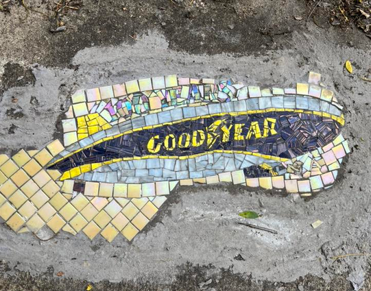 Color image of an urban street art placement in Akron, Summit County, Ohio, USA. Image shows a small mosaic art installation placed into and filling a hole in the sidewalk just outside the International Headquarters of Goodyear, celebrating the 100th anniversary of commercial airship operations by Goodyear in 1925. The mosaic is in shades of gray, blue, and yellow, and the tiles and pieces in the mosaic create the image of a modern Goodyear airship, with the words "100 Years" above the airship.