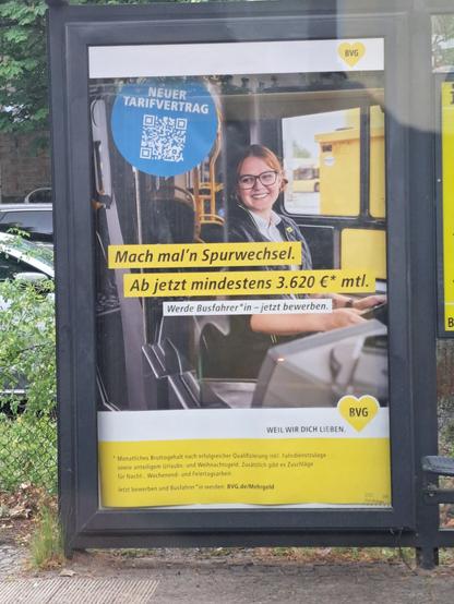 Ein Werbeplakat der BVG an einer Bushaltestelle. Auf dem Plakat ist eine weiblich gelesene Person am Steuer eines Busses abgebildet. Auf einem blauen Sticker steht "neuer Tarifvertrag" mit einem QR-Code dazu und "Mach mal'n Spurwechsel. Ab jetzt mindestens 3.620€ mtl. - Werde Busfahrer*in - jetzt bewerben."