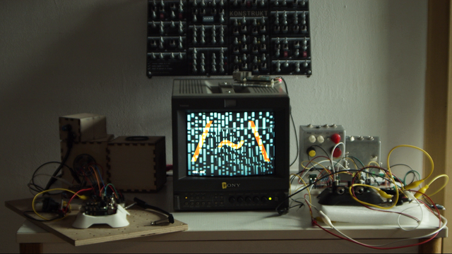 A photo of a table with a turned on CRT in the center of it. Around the CRT there's various electronics projects at different stages of readiness: breadboards, diy pedals, diy speakers, a bunch of alligator clips binding together who knows what. Quite a messy picture.
