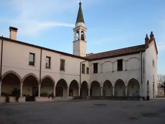 The image depicts a historic architectural structure with a courtyard. It features a building that has arches and a clock tower with a pointed roof. The surrounding atmosphere appears calm and serene, indicative of an older style of architecture, possibly a monastery or a historical town square. There are no people present in the image.

Image Credits: Wikimedia / Mariline di Wikipedia in italiano / Public domain