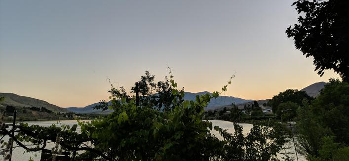 A photo looking west 59 seconds after sunset on June 9, 2025. The sun has disappeared behind a hill to the left of the view an hour previously. There is a grape vine reaching into the sky in the middle of the photo that is obscuring part of a distant mountain. There is a reservoir of water behind the grapevine.