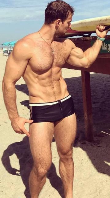 A muscular white man wearing black swimming trunks is flexing his bicep while holding the surfboard