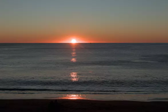 The sea with a beautiful sunset. The sky in the horizon is red as if the sky was on fire. It dissolves into a clear and cold blue.
The sea is very very calm with almost no waves. The sand mimics the color of the approaching night, as well as the dark blue of the water.
Reflections from the sun hit and bounce from the uneven surface of the sea.