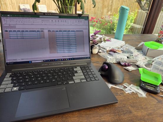 A laptop on a table showing an Excel spreadsheet, to the side is a mouse, a weighing scales, and bags of jump rings