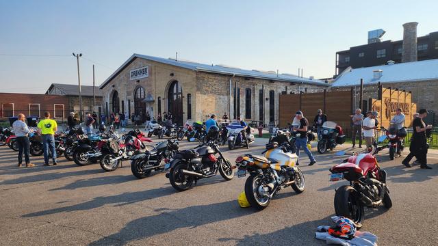 The image depicts a gathering of motorcyclists in an outdoor setting, likely at a motorcycle event. In the foreground, several motorcycles are parked, showcasing a variety of models and colors, including black, red, white, and yellow. The motorcycles are arranged in rows, with some facing the camera and others angled away. The ground is paved, and shadows cast by the motorcycles and people suggest it is either early morning or late afternoon.

In the background, a large brick building with arched windows and a sign reading "DREKKER" is visible. The building appears to be a repurposed industrial structure, possibly a former train station or warehouse. Several people are standing and walking around, some engaged in conversation, while others are inspecting the motorcycles.
