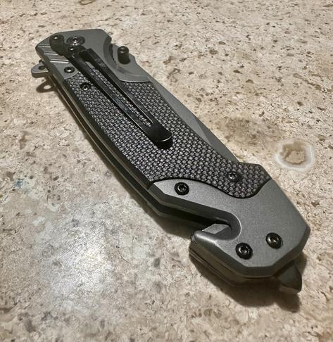 A gray folding knife with a textured black grip, a seatbelt cutting hook and a security crystal breaker is lying on a speckled surface.