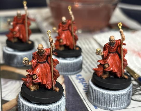 Four 28mm miniatures of necromancers. They are bald, bearded men in red robes holding a skull in one hand and a gold-tipped staff in the other.