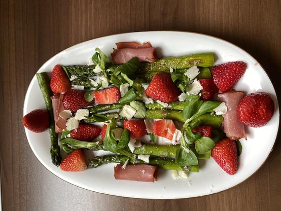 Das Bild zeigt einen Teller mit Salat.  Gegrillte Spargelstangen sind mit Erdbeeren, Feldsalat, Parmesanraspeln und dünnen Scheiben von Schinken (wahrscheinlich Prosciutto) angerichtet. Die Farben sind frisch und kontrastierend – das Grün des Spargels, das Rot der Erdbeeren und das Rosa des Schinkens.  Die Anordnung der Zutaten ist nicht streng regelmäßig, sondern eher locker und modern.