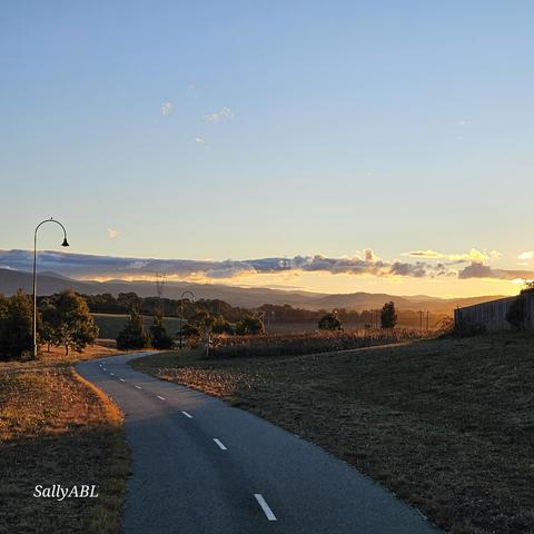 Wide frame colour photo of open bush land. Through the centre of the image is a walking path winding ahead in the direction of a low mountain range on the horizon. From the right is the warm glow of a sunset crossing the top half of the image.  The sunset is reflected on a thin line of clouds parallel to the mountains.