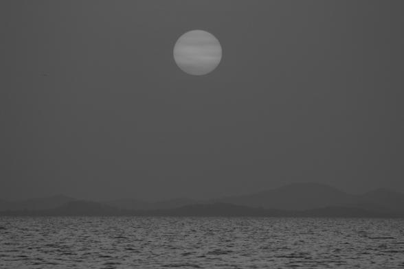 das bild zeigt eine schwarz-weiß-aufnahme eines sonnenuntergangs über einem meer. die sonne ist als runde scheibe am himmel zu sehen, während der rest des himmels und die umliegende landschaft in verschiedenen grautönen gehalten sind. im hintergrund sind schwach erkennbar, durch nebel oder schlechtes wetter verdeckte, bergketten zu sehen. das meer ist recht ruhig und die oberfläche wirkt leicht gewellt. insgesamt vermittelt das bild eine ruhige, melancholische stimmung.