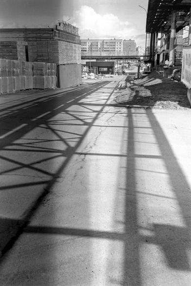 The monochrome photo shows shadows on the asphalt from metal structures. To the left are warehouses. Multistory buildings can be seen in the distance.