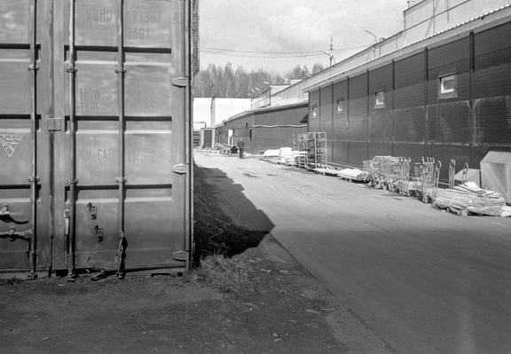 In monochrome photo, part of a warehouse in the foreground. In the far background are other warehouses and a smoking worker.