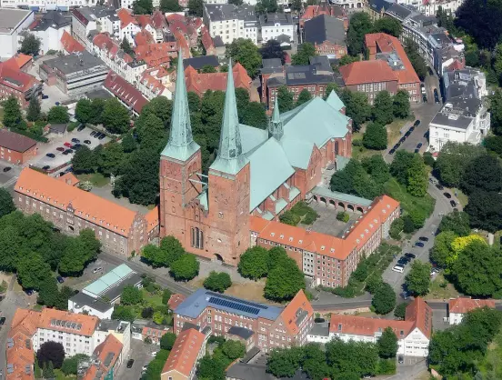 The image depicts an aerial view of a large church or cathedral with distinctive green spires, surrounded by buildings and trees. The architecture appears to be historical, characterized by brick construction and a prominent roof. The surrounding area includes various rooftops, likely residential or commercial buildings, interspersed with greenery.

Image Credits: Wikimedia / Carsten Steger / CC BY-SA 4.0