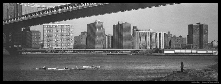 A person stands on a rocky shore with the Manhattan skyline visible across the East River.  A group of four kayaks paddles in the river between them, beneath a large bridge.
