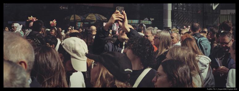 A person stands with their phone raised above their head to take a picture of something in a different direction from the prevailing motion of a large crowd around them.