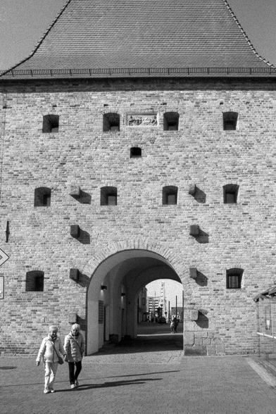 In the monochrome landscape of Rostock, Germany, an elderly couple walks by the imposing SteinTor. The large archway, with its aged bricks and shadowy alcoves, suggests a rich history spanning centuries. In the background, distant buildings appear with softened outlines, a result of the photographic technique employed. However, a modern street sign interrupts this historical scene.