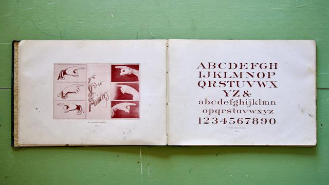 Old book opened out and laid flat to show a spread that has a number of pointing hand graphics and photos on the left sign and a Roman alphabet on the right page.