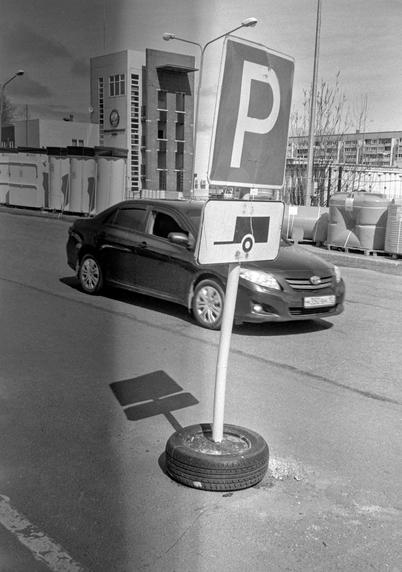Monochrome photo shows a road sign and its shadow. A car is passing by.