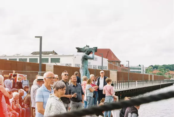 Eine Traube von Menschen steht an der Hafenkante in Flensburg. Die meisten von ihnen schauen in eine Richtung, irgendwo rechts oben vom Bild. Hinter den Menschen die Statue des Christophorus, der ein Schiff auf seinen Schultern trägt.