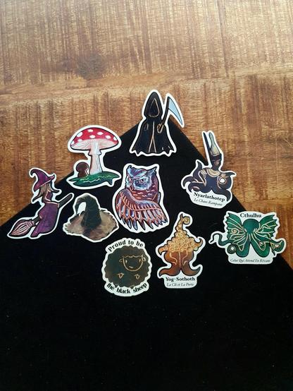 Lots of stickers in front of a black and wood background: Death, a red mushroom, a brown owl, a purple witch on her broom, a witch hat, a black sheep and Lovecraft inspired Great ones Chtulhu, Nyarlathotep and Yog-Sothoth.