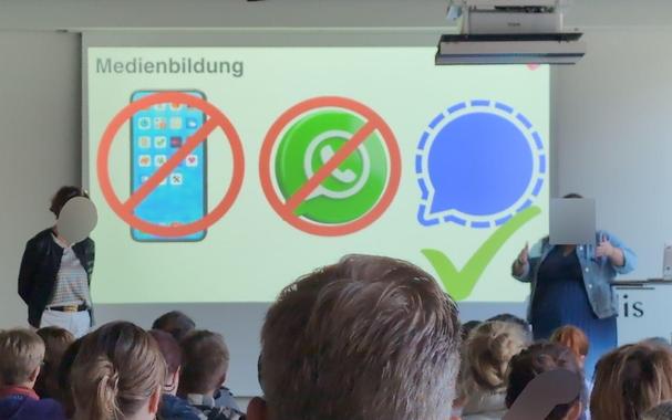 Blick auf das Podium eines Elternabends mit einer Folie, darauf mit Überschrift "Medienbildung" ein Smartphone und ein Whatsapp-Logo jeweils durchgestrichen, und ein Signal-Logo mit grünem Häkchen.