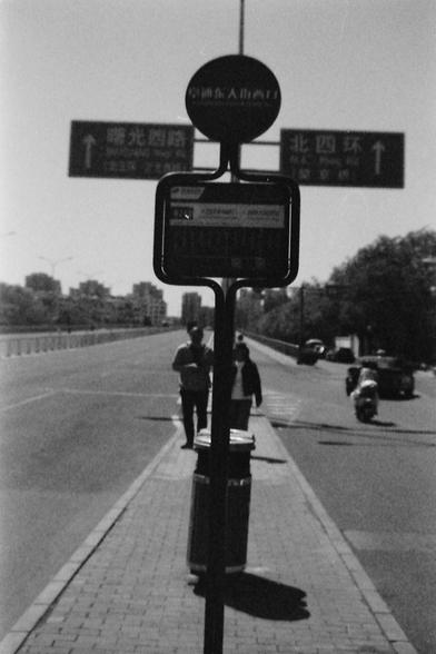 Ilford FP4 E.I. 64

English alt text: A black and white photograph of a street scene with a sidewalk and a bus stop sign in the foreground. Two people are walking on the sidewalk, and a motorbike is on the road. In the background, buildings and trees are visible, along with two large road signs with arrows and Chinese characters.

Chinese alt text: 一张黑白照片，展示了一条街道景象，前景是人行道和公交车站牌。两个人在在人行道上行走，路上有一辆摩托车。背景中可以看到建筑物、树木，以及带有箭头和汉字的两个大型路牌。