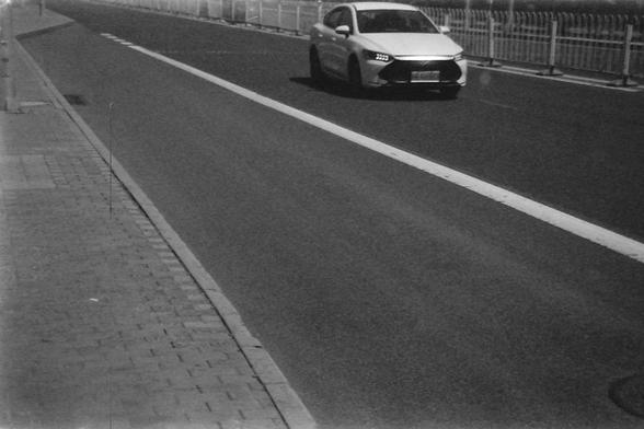 Ilford FP4 E.I. 64

English alt text: A grayscale image of a road with a white car driving toward the camera. The road is marked with a white lane line and has a brick-paved sidewalk on the left side. A fence runs parallel to the road in the background.

Chinese alt text: 一张灰度图像，展示了一条道路，一辆白色汽车正朝着镜头驶来。道路上有白色车道线，左侧是铺有砖块的人行道。背景中有一条与道路平行的围栏。