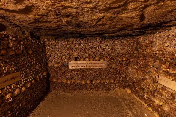 The image depicts a subterranean area, likely a skull and bone ossuary or catacomb, with walls lined with human bones. This setting often serves as a burial site or a place to store skeletal remains, typically seen in historical or archaeological contexts. The arrangement of the bones suggests an intentional display or organization. There are also signs in the image, likely providing information about the site.

Image Credits: Wikimedia / Diego Delso / CC BY-SA 4.0