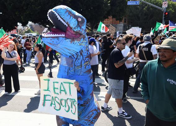 A person in a dinosaur costume holding a sign which reads:
I EAT FASCISTS.