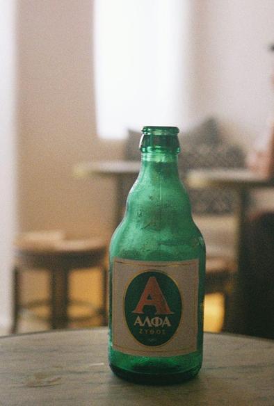 Color photograph of a green beer bottle sitting on a wooden table. The beer label has a large red A on a green oval background with ΑΛΦΑ written below.