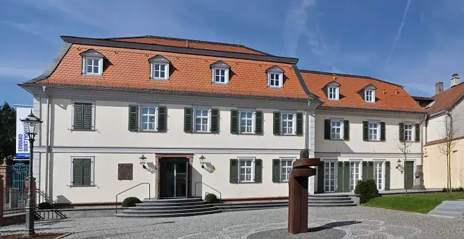This image appears to show a building with a distinct architectural style, featuring a symmetrical façade, a sloping roof with multiple dormer windows, and shutters on the windows. There's also a sculpture or a modern artistic structure in the foreground, along with a paved area. The setting seems to suggest a formal or institutional purpose, possibly a museum or cultural center, but without specific context, I cannot identify it further.

Image Credits: Wikimedia / Eva K. / GFDL 1.2