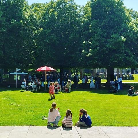 Lawn with a ice cream cart under trees. A  parasol offers shade to a group of sitting people on chairs. In the foreground three girls eating their ice cream. | Grasveld met een ijscokar onder de bomen. Een parasol biedt schaduw aan een groep mensen die op stoelen zitten. Op de voorgrond eten drie meisjes hun ijsje.