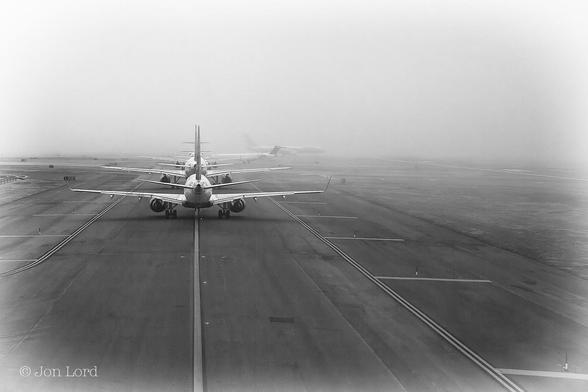 A Low Contrast Black And White Landscape Photo In Landscape Format. One third Of The Way From The Lower Left Corner Is A Thick Line Stretching Away From The Camera. The Line Is Painted On A Wide Strip Of Tarmac Perhaps Fifty Or Sixty Metres Wide. Either Side Of The Painted Line Are A Two Pairs Of Double Lines, On Either Side Of The Central One And Perhaps Ten Metres Offset. To The Right Of The Centre Line Are Lights Partially Inset Into The Tarmac. Either Side Of Tarmac Is An Area Of Short Grass. Surrounding This Scene Is A Body Of Thin Fog Or Mist Obscuring The Sky And Anything Beyond One km. About Twenty Metres Ahead Of The Camera And Facing Away Is A Line Of Six Or More Small And Medium Sized Jet Airliners.
The Location: JFK Airport On Long Island, New York (2011).