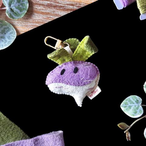 Picture of a handmade felt turnip bag charm photographed on a black and wooden background, surrounded with decorative plants and matching felt and threads. 
The bag charm is purple and ivory with three leaves coming out of the top, curling on themselves slightly. An ivory ribbon is attached to the middle of them, linking the turnip to a golden lobster clasp. 
The turnip has two curious eyes embroidered in black.