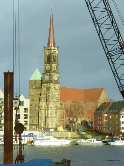 The image features a prominent church or cathedral with a tall steeple, set against a backdrop of buildings and a body of water. The structure appears to have a mix of architectural styles, with a clock visible on the tower and distinct rooftops on the adjacent buildings. The scene suggests a waterfront location, possibly indicating a port or riverside area, with boats moored nearby. The weather looks cloudy, adding a dramatic effect to the scene.

Image Credits: Wikimedia / Ulamm (Diskussion) / CC BY-SA 4.0