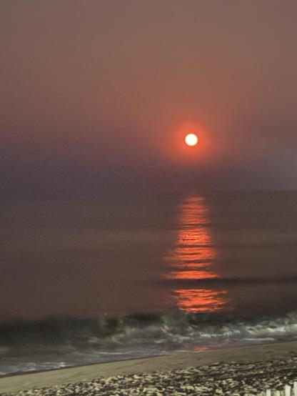 Strawberry Full moon  rises over the sea.  It is dark, and the moon is an intense red, it's the same color reflecting off the calm sea.  Small waves break on the beach in the foreground.