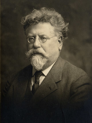 A middle-aged man stands alone in a studio setting with a muted backdrop. He wears a formal suit and tie, typical of his era. His expression is neutral, and the composition directs attention solely to him. The photograph is of Rudolf Rocker, a prominent German anarchist writer and activist.