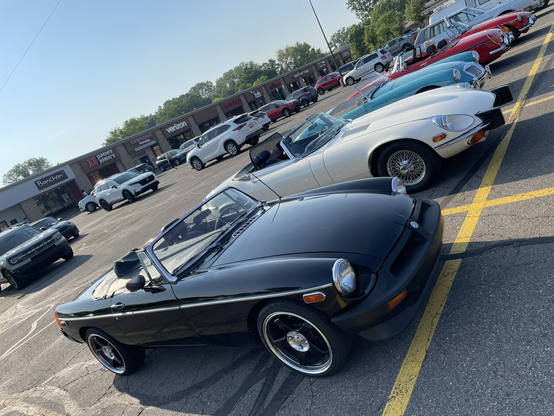 Photograph tilted down and to the left of a black rubber-bumper 1976 MGB next to a white 1974 Jaguar XKE (aka E-Type) in a strip mall parking lot. Several other small British cars can be seen behind them.